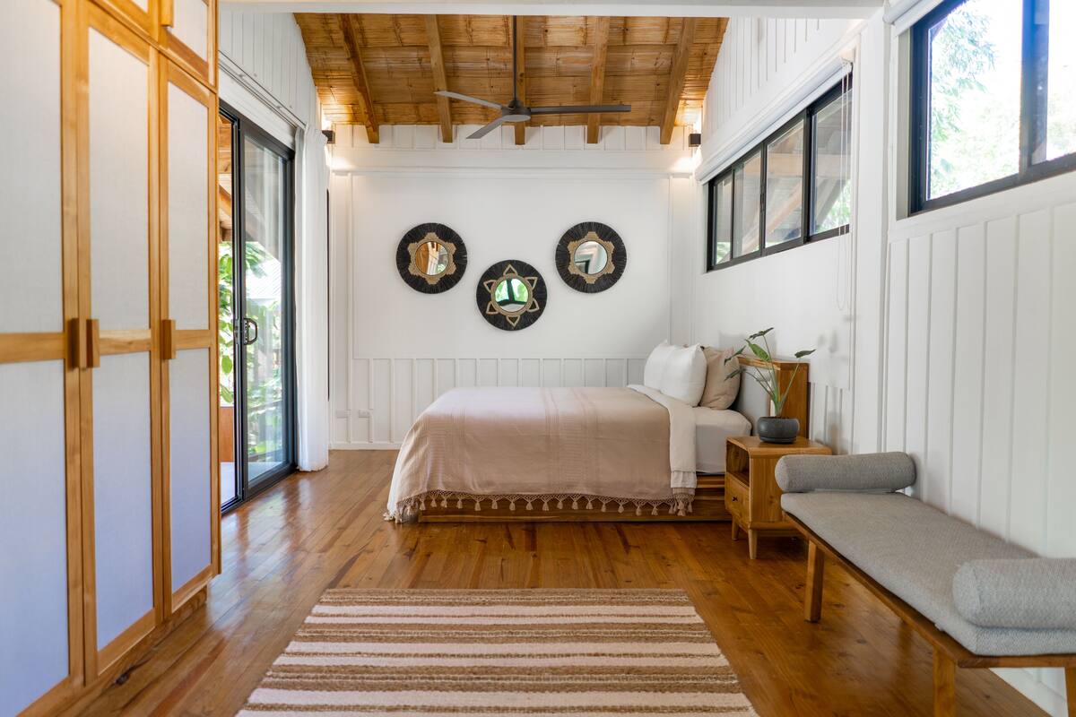 A bedroom with a bed, a bench, a bedside table with a plant, and three wall mirrors.