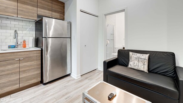 A compact kitchen with a stainless steel refrigerator, wooden cabinets, a black leather sofa, and a small table. - River West, Third Floor Chicago by 747 Lofts (Chicago)