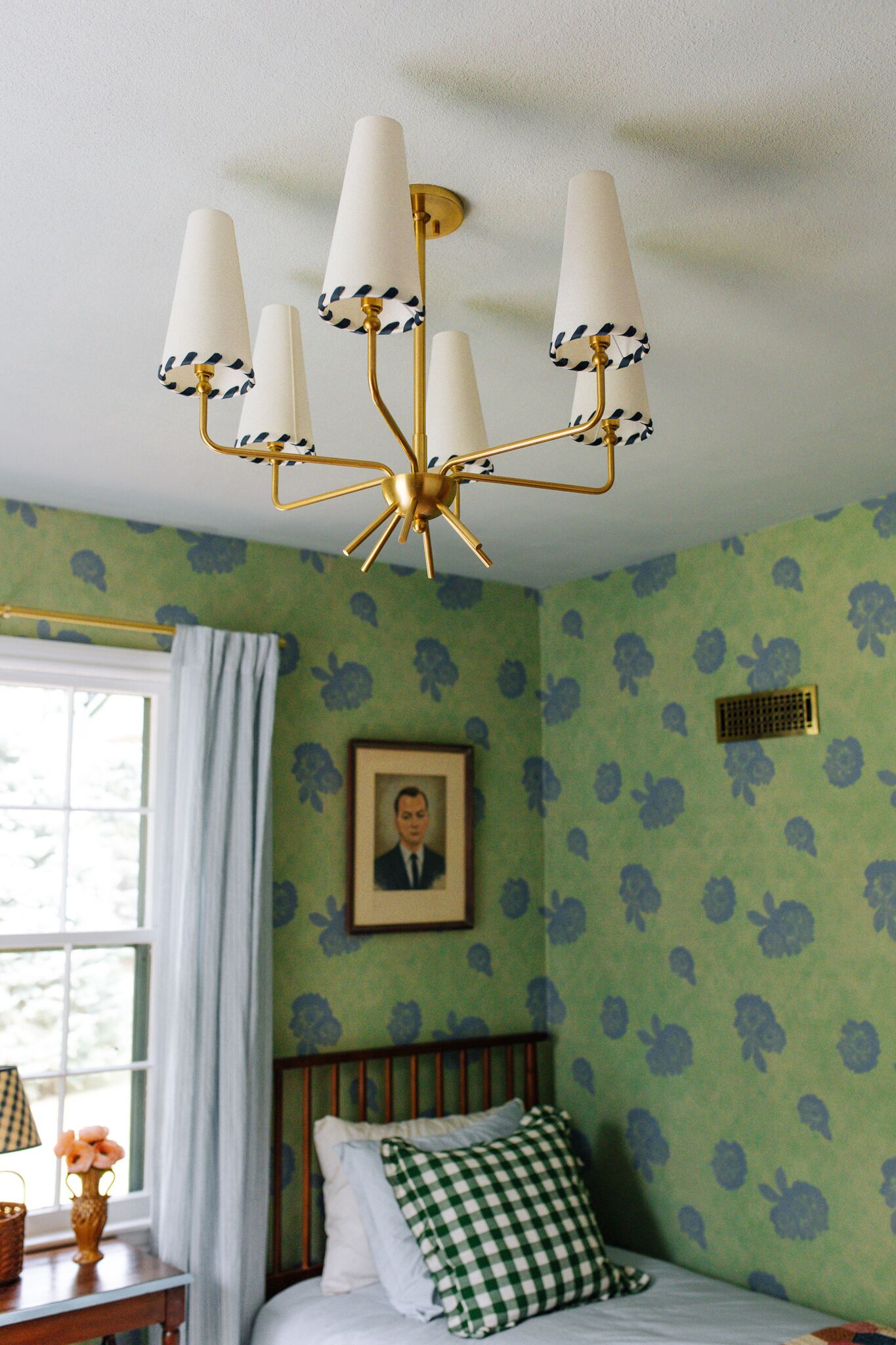 Une chambre à coucher avec un papier peint à motifs floraux verts, un lit avec des oreillers à carreaux blancs et verts, un portrait encadré accroché au mur et un lustre doté de plusieurs abat-jours.