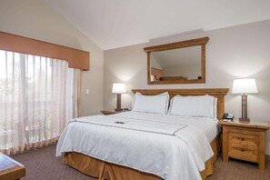 Quarto de hotel com uma cama grande, duas mesas de cabeceira com abajures, uma cabeceira de madeira, um espelho sobre a cama e cortinas finas na janela.