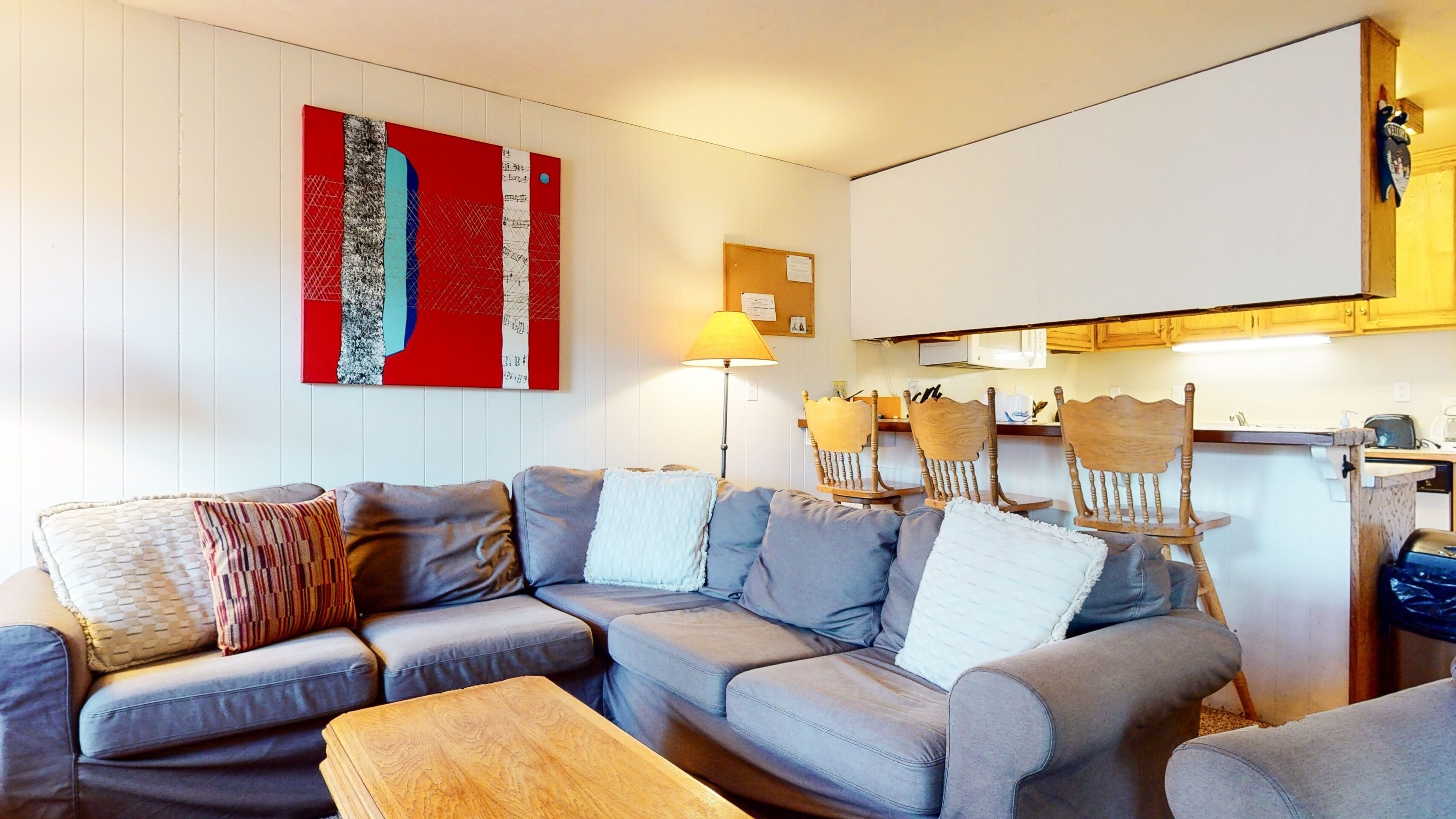 A living room with a grey sectional sofa, wooden coffee table, and a wall-mounted TV. There are wooden chairs and a lamp in the room.
