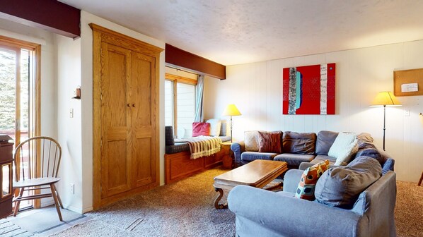 Una sala de estar acogedora con una puerta de madera, un sofá, una silla, una mesa y una ventana con cortinas.