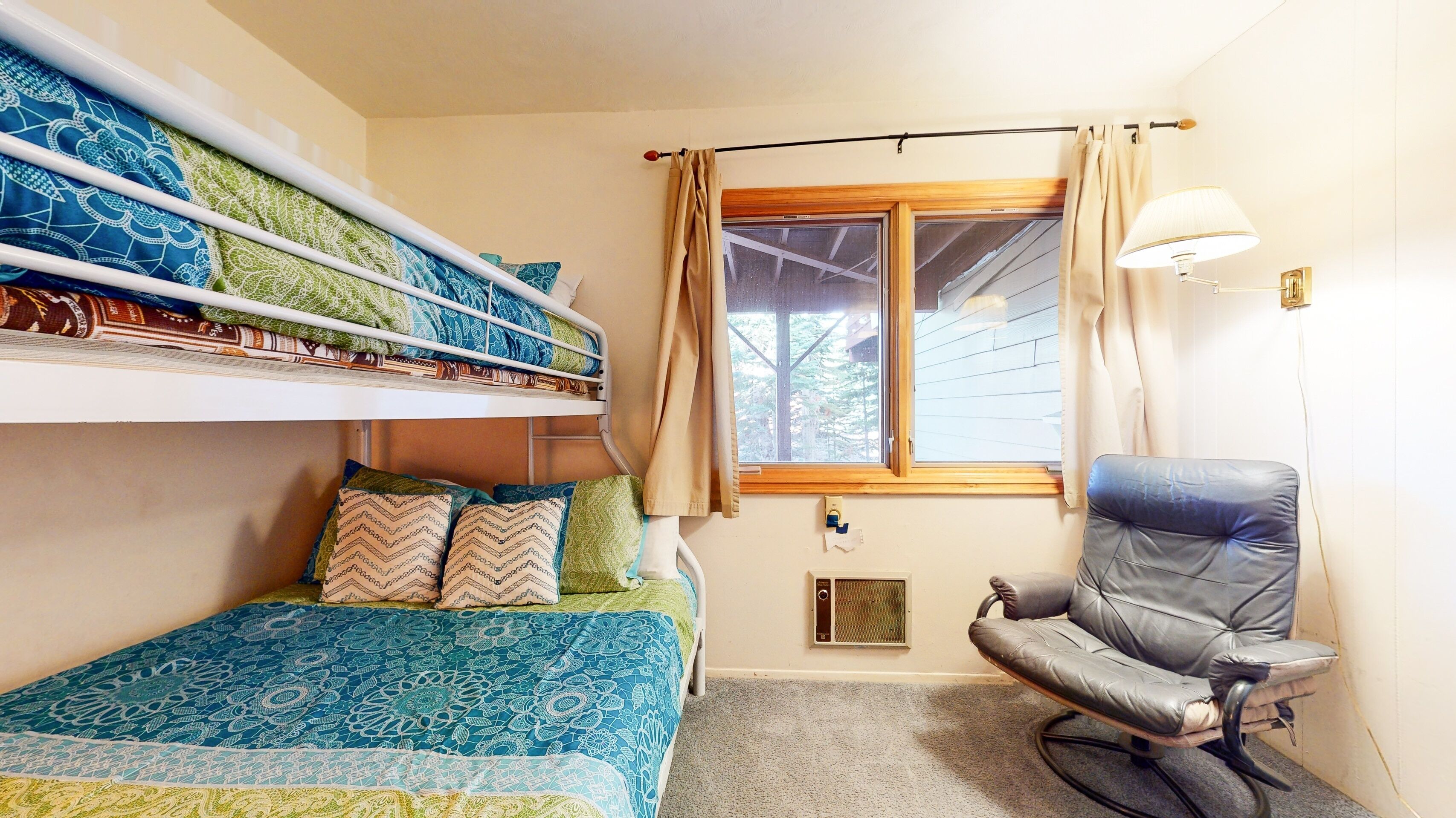 Una habitació amb lliteres, una cadira, una finestra amb cortines i un calefactor fixat a la paret.