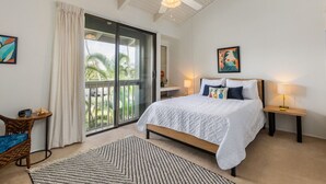 Une chambre avec un grand lit, un ventilateur de plafond, une vue sur les palmiers à travers une porte-fenêtre coulissante et une petite table avec une chaise.