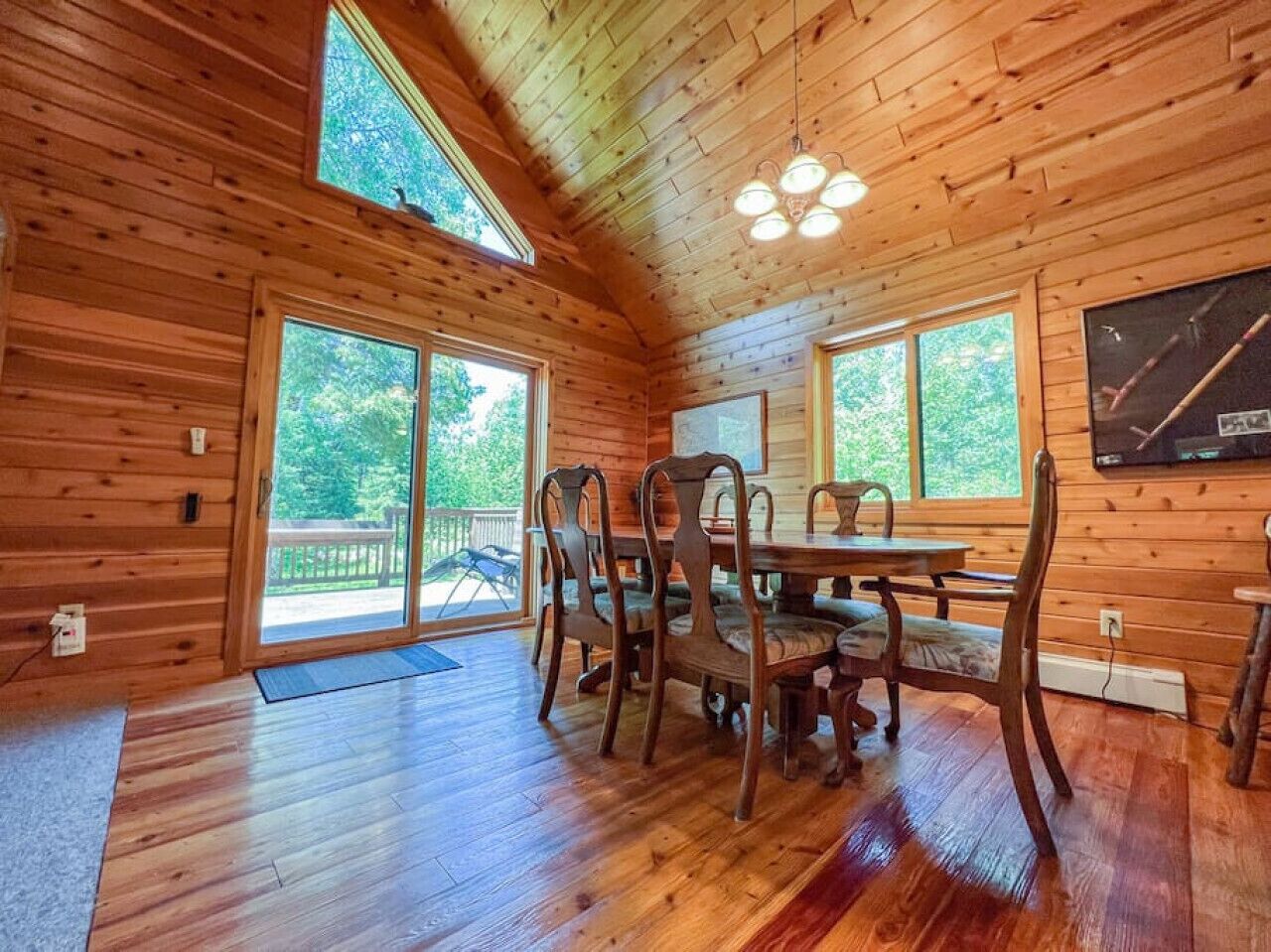 A wooden cabin interior with a dining table and chairs, large windows, and a ceiling light fixture.
