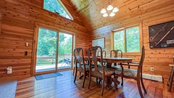 A wooden cabin interior with a dining table and chairs, large windows, and a ceiling light fixture.