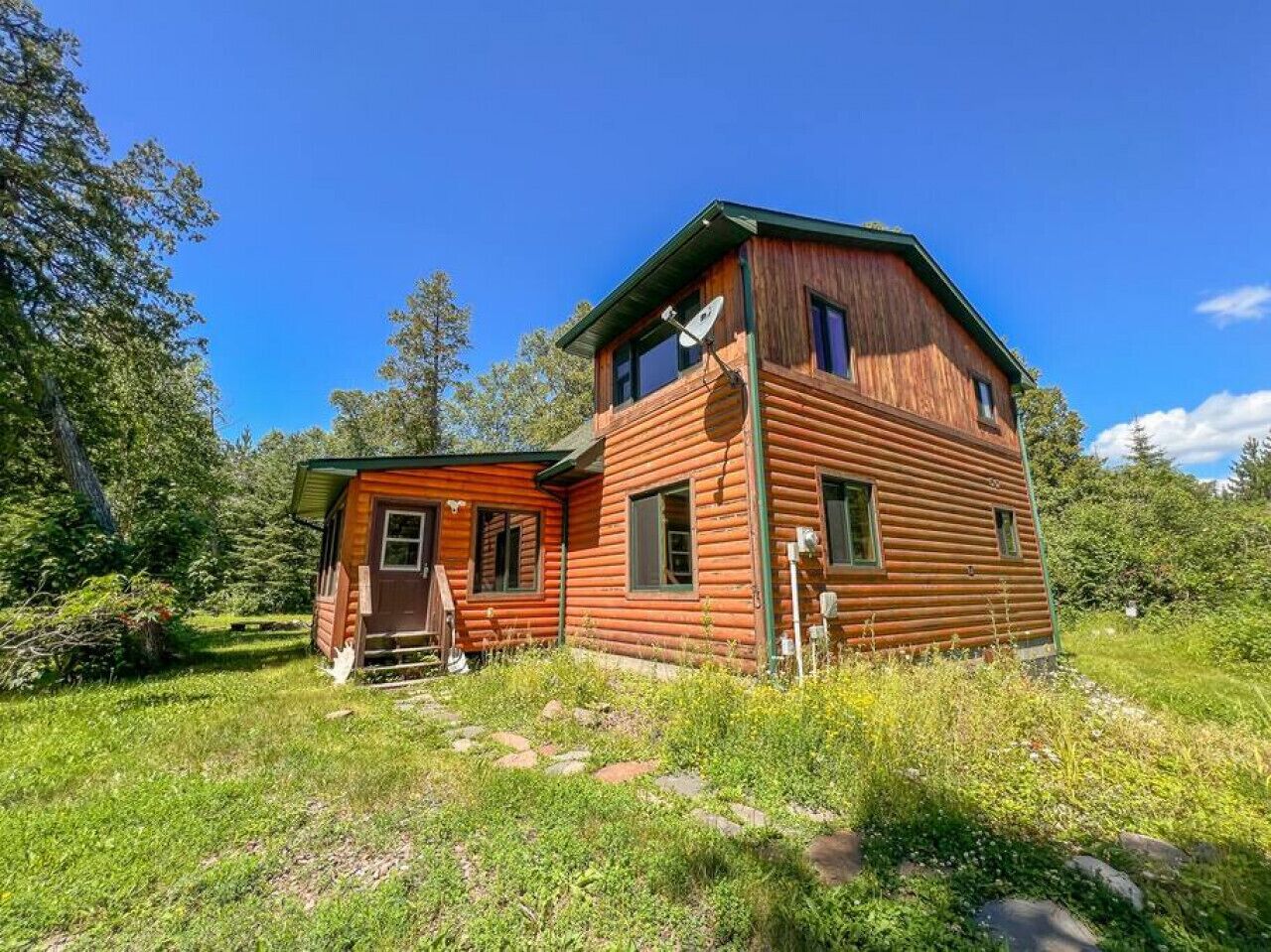 A wooden cabin with a small attached shed, surrounded by lush greenery and trees.