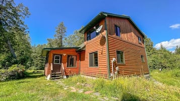 A wooden cabin with a small attached shed, surrounded by lush greenery and trees.
