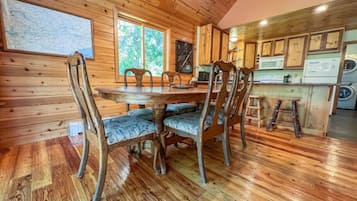 A wooden interior with a dining table and chairs, a kitchen area with appliances, and a large window providing natural light.