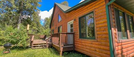 A log cabin with a deck, a grill, and a forested backdrop.