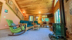 Ein gemütliches Blockhaus-Interieur mit Holzbalkendecke, einem grünen Schaukelstuhl, einer Couch, einem kleinen Tisch und einem Kamin.
