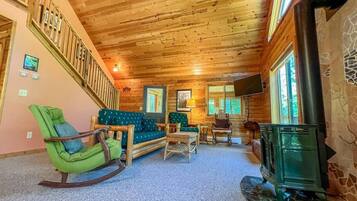 A cozy cabin interior with a wooden ceiling, a green rocking chair, a sofa, a small table, and a fireplace.
