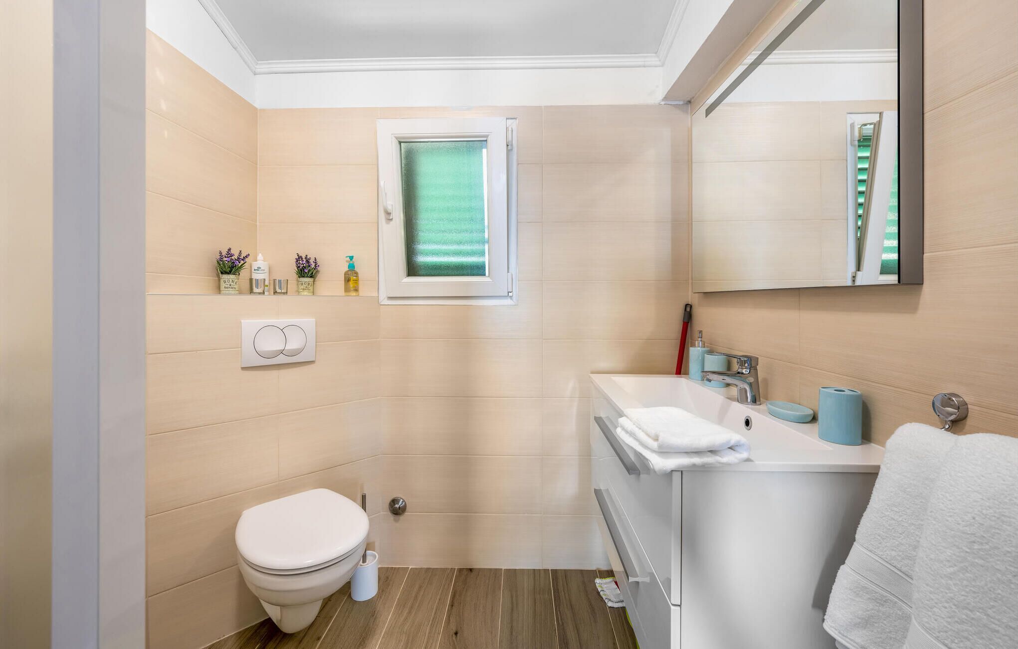 A modern bathroom with a toilet, sink, and mirror. There is a window with a view of greenery, and toiletries are placed on a shelf.
