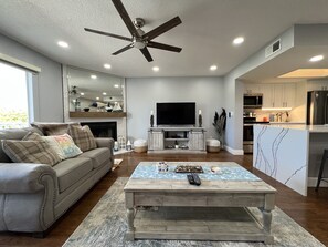 A spacious living room with a large TV, a ceiling fan, and a kitchen area with modern appliances.