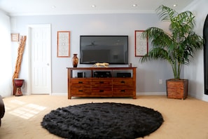 Una sala de estar con un televisor de pantalla plana grande, un mueble de madera para equipo de entretenimiento, un tapete negro y mullido, una planta en maceta y decoración en la pared.