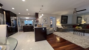 A modern kitchen with dark cabinetry and a central island, a living area with a grey sofa and a coffee table, and a dining area with a glass table. - Mai Villa-Hampton - Where good times happen! (Hampton)