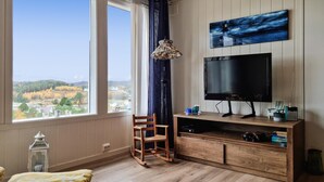 A room with a large window, a TV, a wooden chair, and a small table with books and a lamp.
