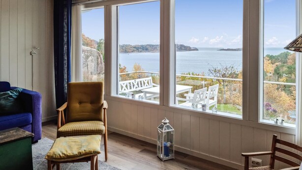 A room with a large window overlooking a lake and autumn foliage. There is a blue sofa, a yellow armchair, and a small table with a lantern.