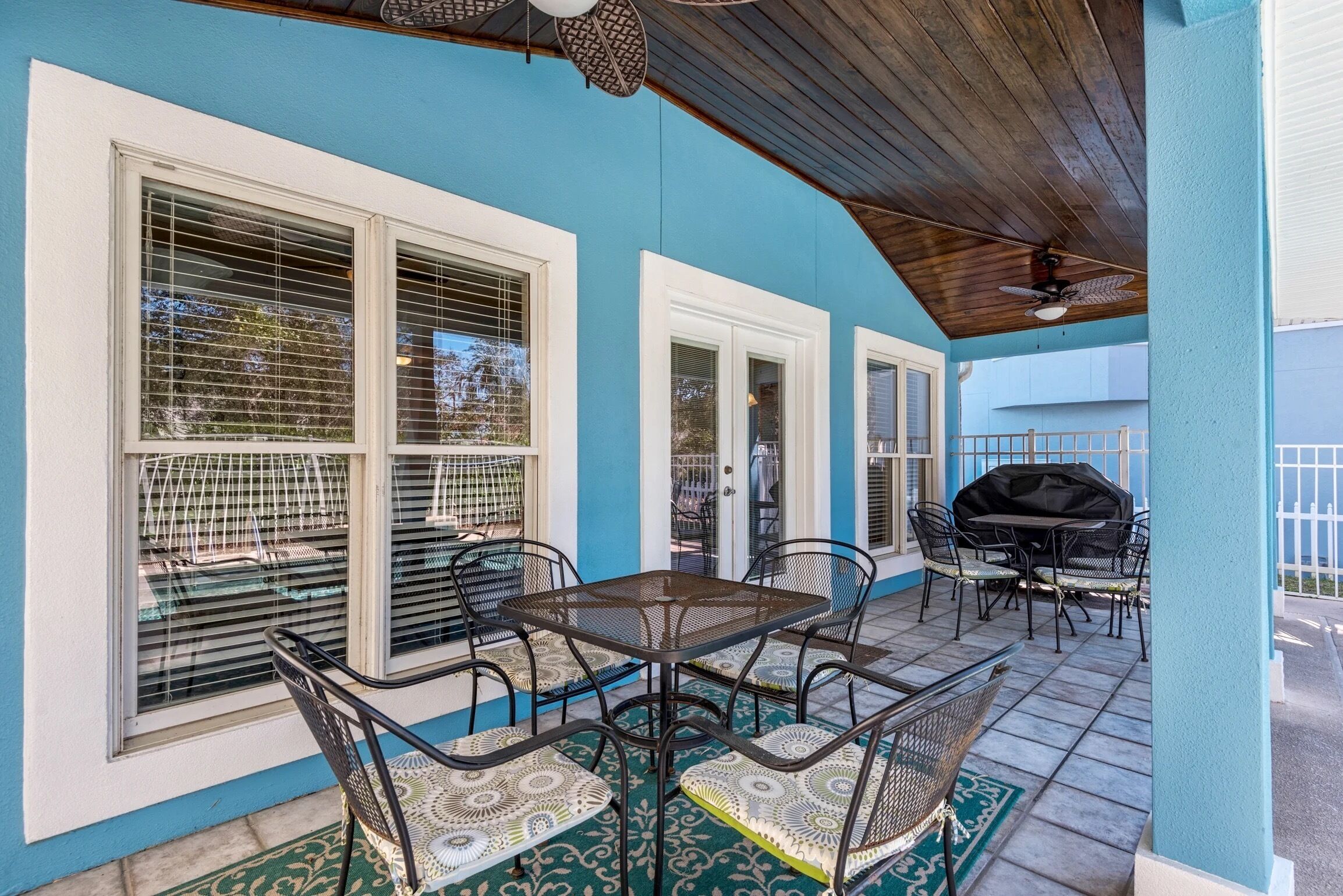 Une terrasse couverte avec une table et des chaises, un barbecue et de grandes fenêtres équipées de stores.