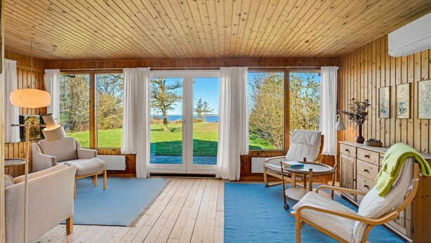 A cozy room with wooden walls and ceiling, large windows offering a view of a grassy area and trees, a sofa, a chair, a small table, and a cabinet. - 3 bedroom cozy home in Spøttrup (Spøttrup)