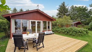 A wooden deck with a table and chairs, a red building with large windows, and a garden with various plants. - 3 bedroom cozy home in Spøttrup (Spøttrup)