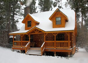A log cabin with a porch and a chimney, surrounded by snow and trees.