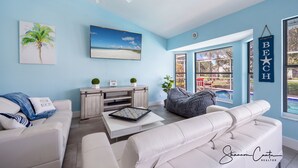 A modern living room with a white couch, a coffee table, and a bean bag. There is a painting of a beach, a clock on the wall, and a sign that reads "BEACH". - The Blue Lagoon of Naples (Naples)