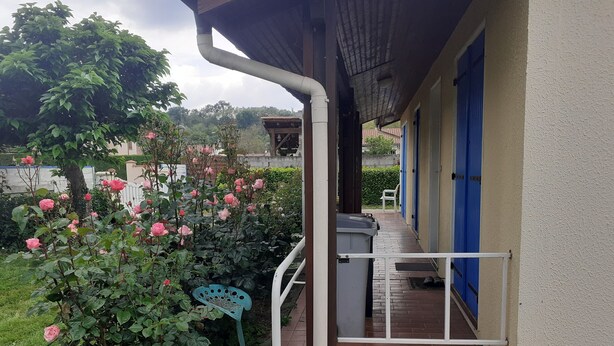 Un espacio al aire libre cubierto con un camino de ladrillos que lleva a un edificio con puertas azules. Hay un jardín con rosas rosas y una silla azul.