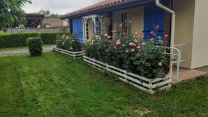 Un edificio con techo de tejas, puertas azules y una valla blanca que rodea un jardín con rosas rosadas.