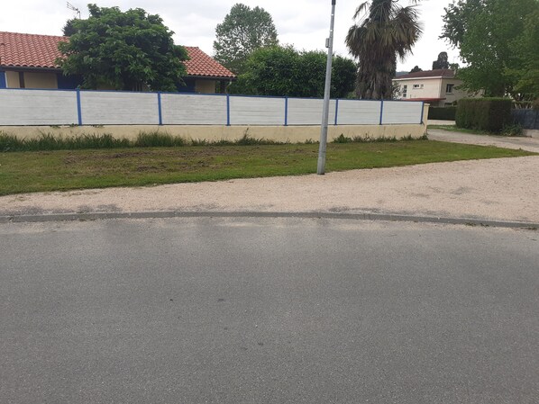 Una carretera pavimentada con una cerca azul y blanca, un área con césped y edificios al fondo con tejados de tejas rojas.