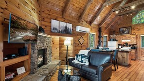 Interior acogedor de una cabaña con chimenea de piedra, sofá de cuero, paredes de madera y una zona de cocina.