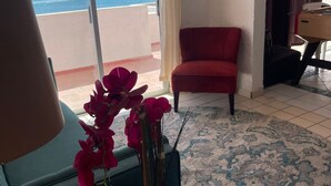 Ein Zimmer mit Blick auf den Ozean, ein roter Sessel, ein Holztisch mit Blumenvase und eine Lampe.