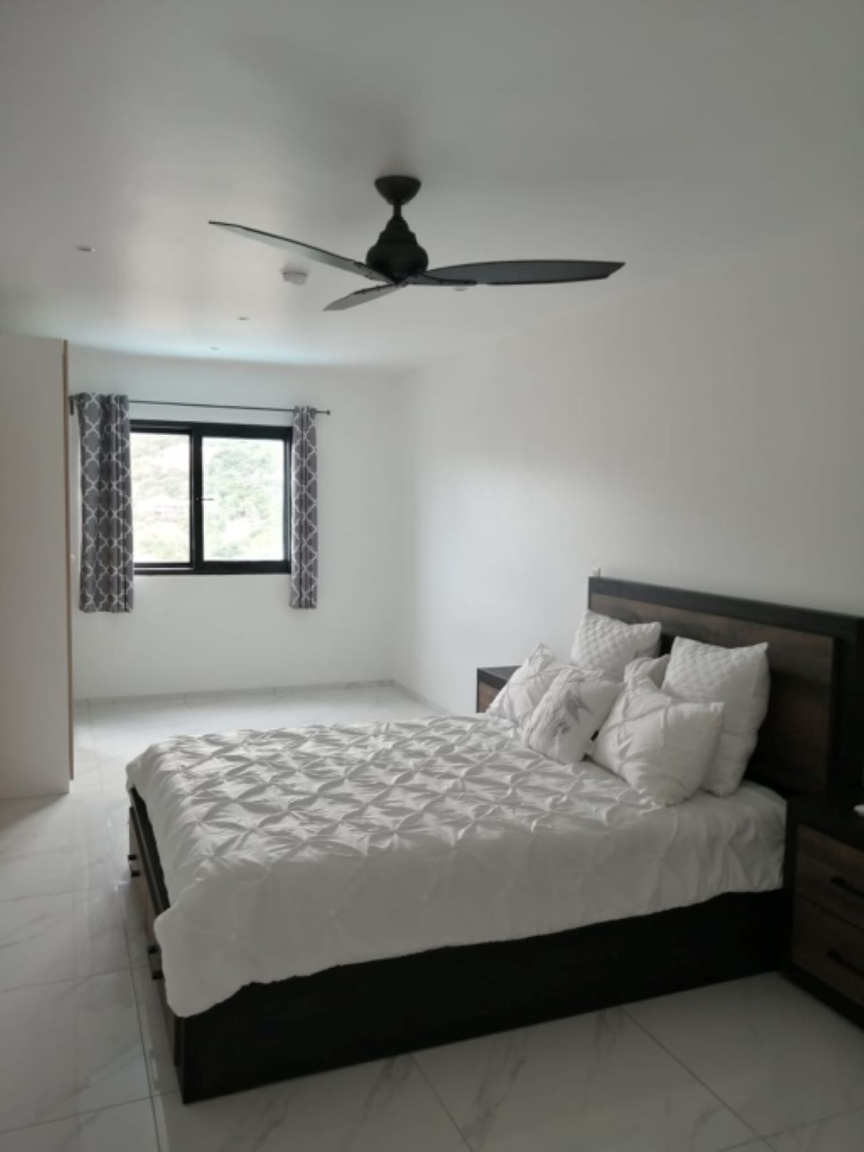 A bedroom with a bed, a ceiling fan, a window with curtains, and a nightstand.