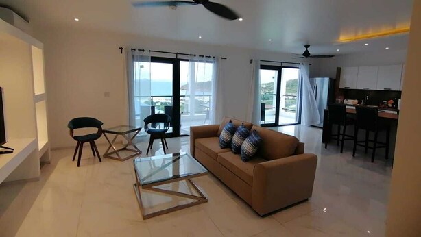 A modern living room with a glass coffee table, a sofa with cushions, a ceiling fan, and large windows offering a view of the outdoors. - Spectacular view of the Caribbean Sea (Sint Maarten)