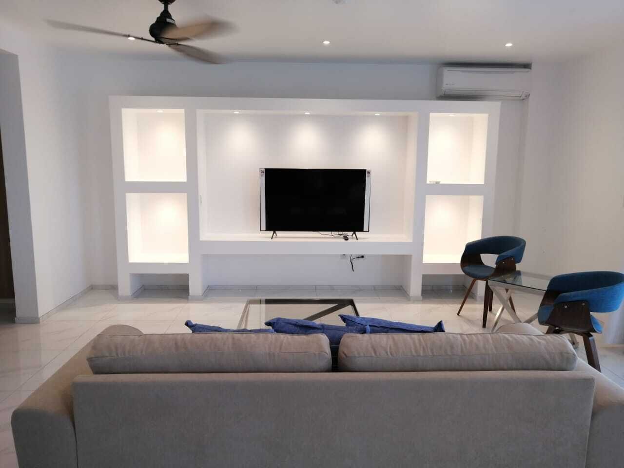 A modern living room with a large flat-screen TV mounted on a wall, a glass coffee table, a beige sofa, and two blue chairs.