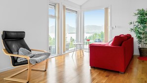 A modern living room with a black leather chair, a red sofa, and a view of a lake through large windows. - 4 bedroom nice home in Jørpeland (Jørpeland)