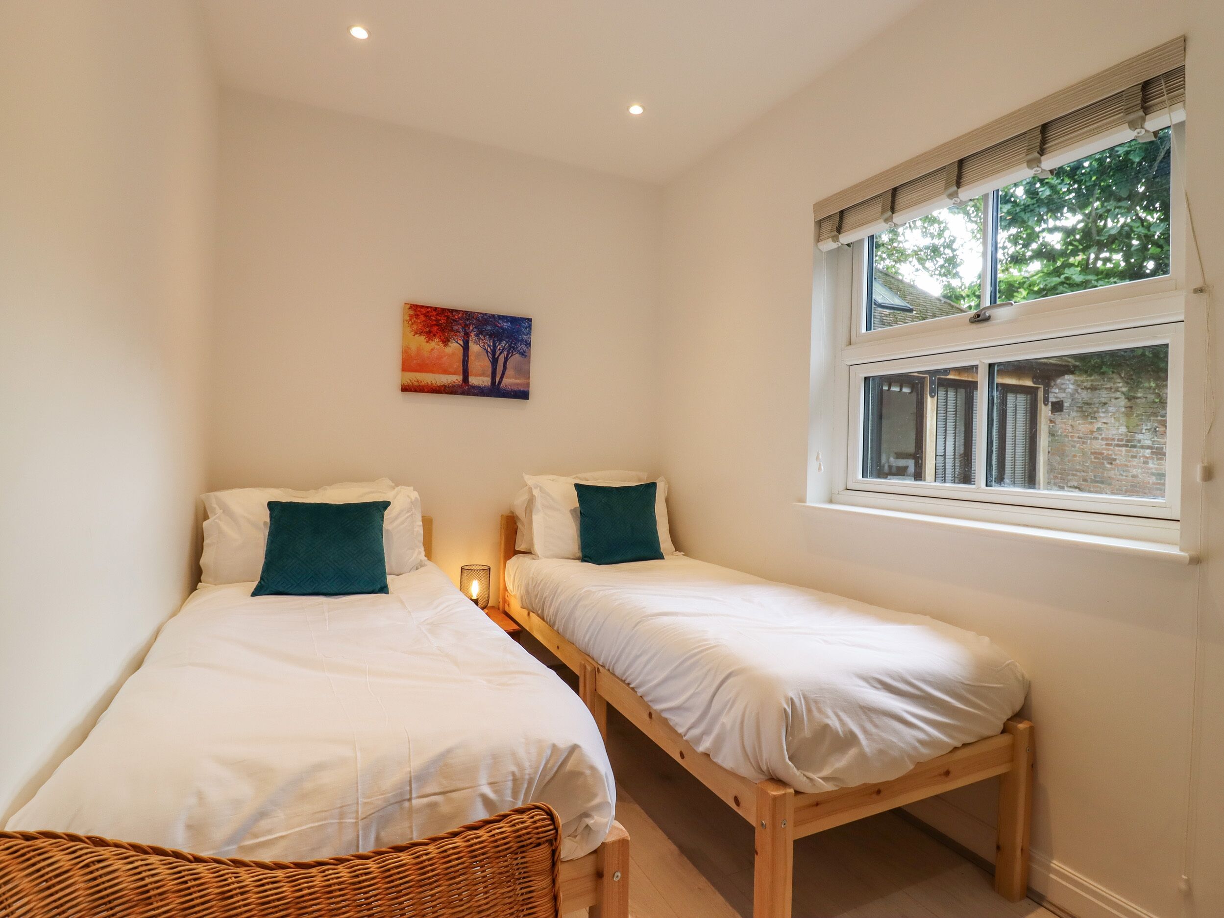 Um quarto com duas camas de solteiro, uma cadeira de madeira, uma mesa de cabeceira com luminária, uma janela com persianas e um quadro na parede.