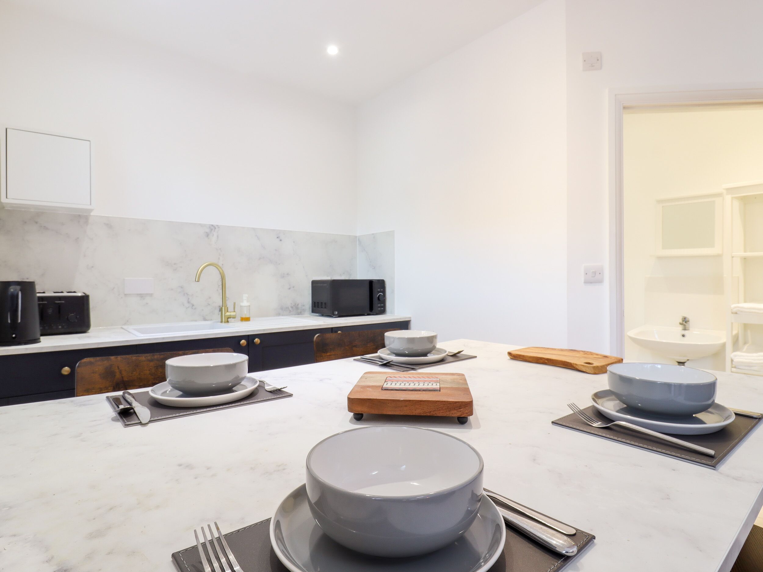 Una cocina moderna con encimera de mármol, un lavabo, un microondas y una zona de comedor con una mesa preparada para dos personas.