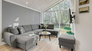 En moderne stue med en grå hjørnesofa, et lille rundt sofabord og en grå lænestol. Der er et stort vindue med udsigt til det fri.
