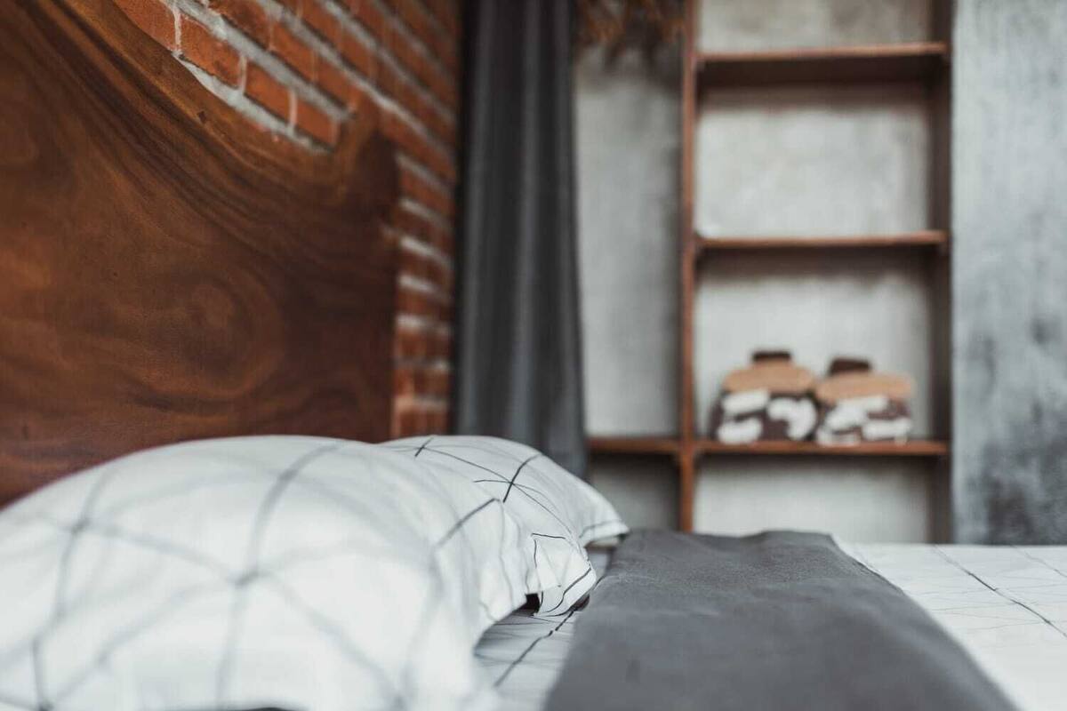 A bedroom with a brick headboard, a white bedspread, and a grey blanket. There is a wooden shelf with shoes in the background.