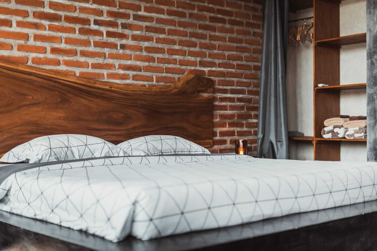 Un lit avec une couvre-lit aux motifs géométriques blancs et gris, une tête de lit en bois et un mur de briques derrière.