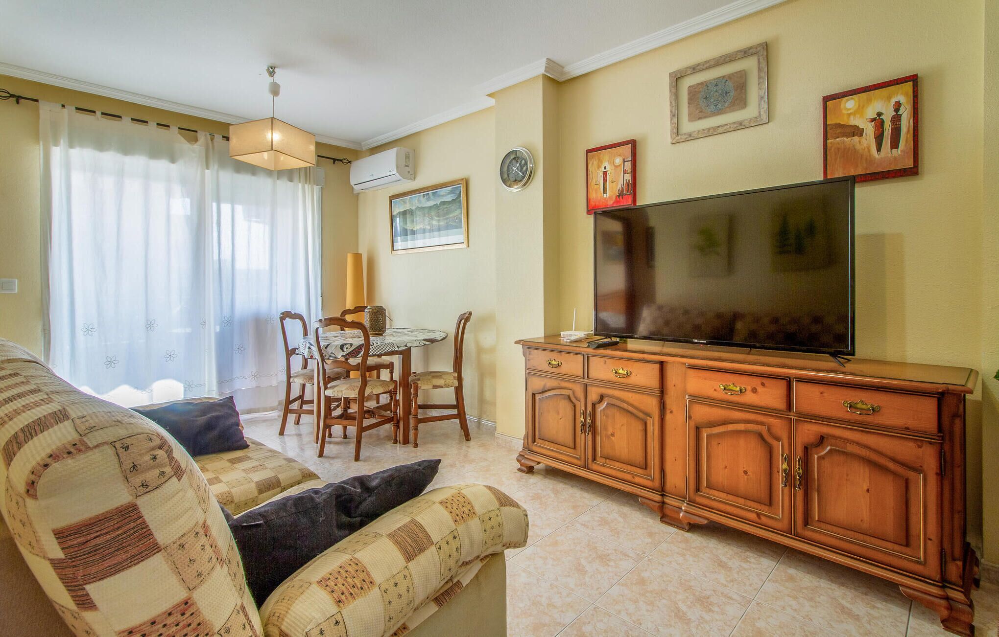 A living room with a sofa, a TV, a wooden cabinet, a dining table with chairs, and a window with curtains.