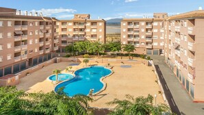 Um edifício residencial com vários andares, piscina cercada por palmeiras e céu claro.