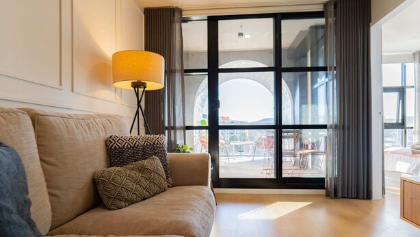 Ein modernes Wohnzimmer mit einer Couch, gemusterten Kissen, einer Stehlampe und einem großen Fenster mit Blick ins Freie.