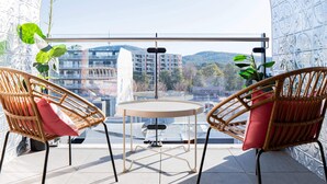 Ein Balkon mit zwei Korbstühlen, einem kleinen Tisch und Blick auf Gebäude und Hügel.