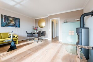 A modern living room with a yellow sofa, a dining table with chairs, a large painting, and a wood-burning stove. - House Sönshörn Whg. 07 (Sylt)