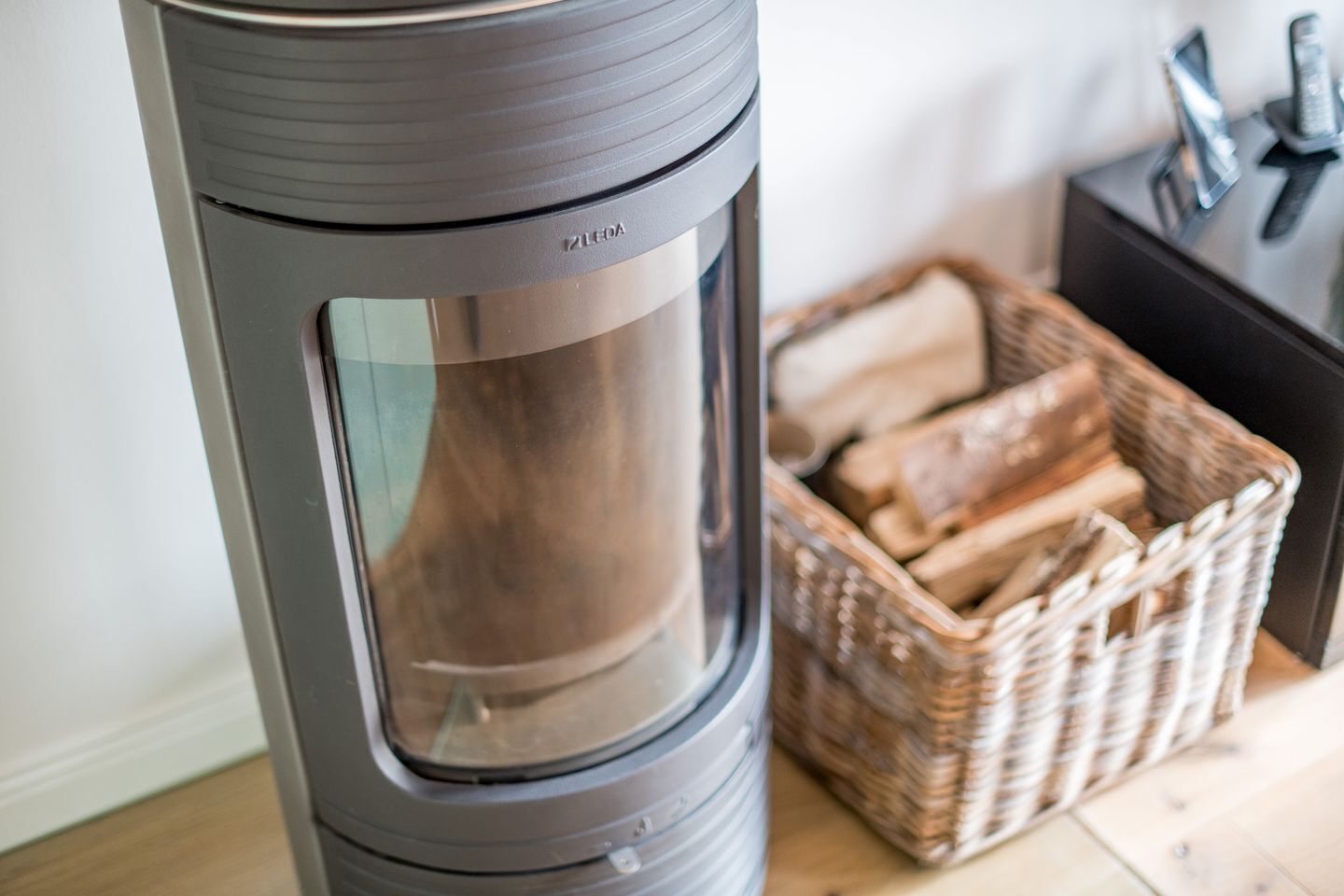 Ein moderner, zylinderförmiger Holzofen mit Glastür, neben einem Korb aus Weidengeflecht, gefüllt mit Brennholz.