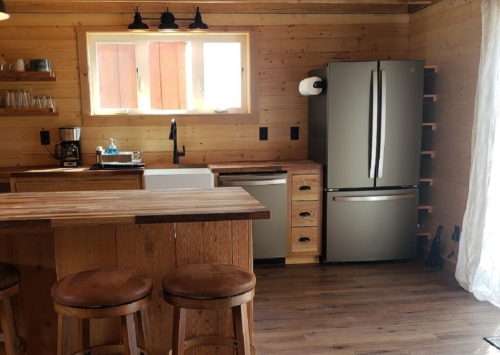 A wooden-paneled kitchen with a large refrigerator, wooden stools, a sink, and a window with blinds.
