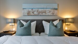 Une chambre d’hôtel avec un lit, deux tables de chevet, deux lampes, un tableau représentant une scène de plage et une tête de lit sombre.