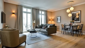 Una sala moderna con sofá, sillón, mesa de comedor y sillas. Hay una lámpara, un televisor y una ventana con cortinas.